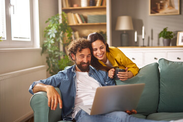 A man and woman are sitting together on a green couch, with the man focused on a laptop while the woman smiles and holds a cup. Sunlight streams into the cozy living room.