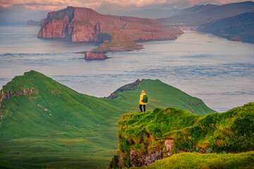 Tourist in yellow jacket admiring of gorgeous mountain landscape, Alaberg cliffs, Faroe Islands, Denmark, Europe. Wonderful summer view of Mykines island with Vagar island on background.