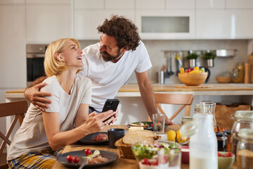 A couple shares a joyful breakfast in their cozy kitchen. They exchange smiles and laughter while enjoying a delicious spread of food on the table, creating a warm atmosphere.