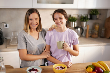 In a bright kitchen, a mother and daughter smile while sharing a breakfast experience. Bowls of...