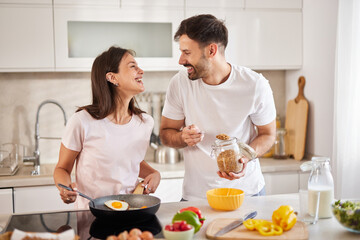 In a bright, modern kitchen, a couple joyfully prepares breakfast. She stirs eggs in a pan while he playfully holds a jar of granola, both smiling and sharing a light-hearted moment.