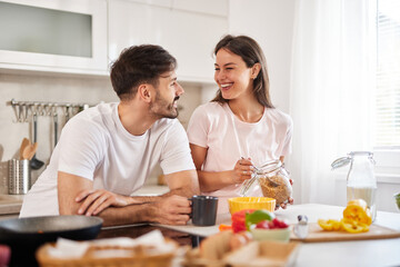 A couple is laughing and sharing a joyful moment while preparing breakfast in their cozy, well-lit kitchen, surrounded by colorful fresh fruits and cooking supplies.