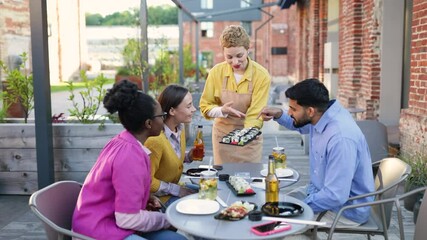 A waitress serves sushi to a group of friends dining outdoors at a restaurant. They are happy.