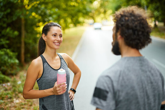 Two runners pause to talk and enjoy hydration during a morning workout on a tree-lined running path, promoting fitness and social interaction in a natural setting. - Powered by Adobe
