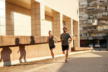 Two runners, a woman and a man, are jogging side by side on a paved path in front of a modern...