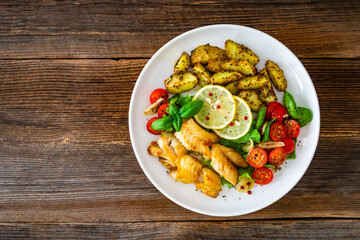 Seared pollock fish, baked potato wedges and tomato salad on wooden table	