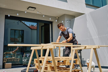 Man working on outdoor carpentry task, measuring wooden board on sawhorses in backyard