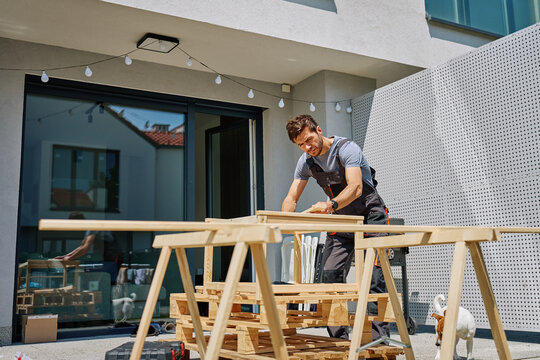 Man working on outdoor carpentry task, measuring wooden board on sawhorses in backyard