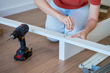 Woman assembling bed frame on wooden floor, using wrench and drill for installation. DIY furniture...