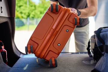 Close up of hand placing orange suitcase into car trunk, preparing for travel or vacation. Inside view of man loading luggage in vehicle