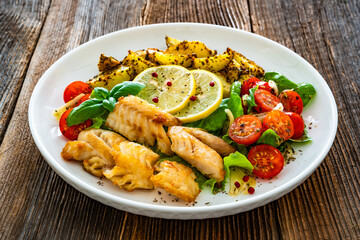 Seared pollock fish, baked potato wedges and tomato salad on wooden table	