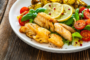 Seared pollock fish, baked potato wedges and tomato salad on wooden table	