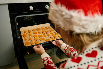 Woman in red festive sweater and Santa hat putting tray of Christmas cookies into oven. Process of...