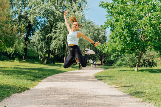 Energetic female leaping high, celebrating fitness amid verdant parkland, embodying vibrant wellness under bright summer sunlight