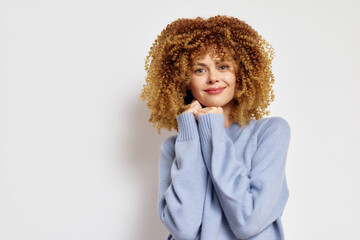 Smiling young woman with curly hair dressed in a light blue sweater against a white background,...