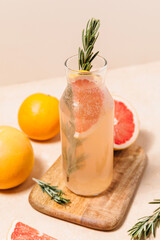 Refreshing grapefruit and rosemary cocktail in a tall glass bottle on a wooden cutting board against a neutral beige background. Summer drink concept, natural organic lemonade recipe for holiday party