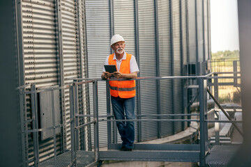Full length portrait of an old senior worker standing near silo and metal construction with tablet in hands.