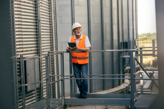 Full length portrait of an old senior worker standing near silo and metal construction with tablet in hands. - Powered by Adobe