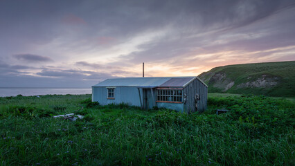 Rustic blue cabin surrounded by lush green grass under a dramatic sky at sunset, showcasing serene...