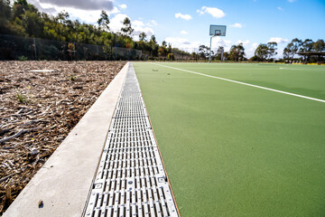 A stormwater drainage grate beside a modern outdoor basketball court in a suburban Australian recreational sports ground. A well-maintained public community infrastructure.