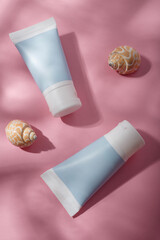 Two blue unbranded cosmetics tubes mockup, seashells on pink background, harsh light with shadows. Beauty product template, advertising banner. Copy space, top view