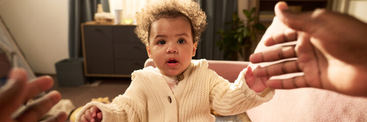 Portrait of baby sitting on sofa reaching out toward hands of Black man indoors, illustrating paternity leave and father child bonding in home environment