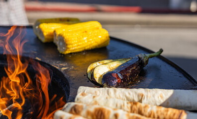 Close-up of corn and meat grilling on a barbecue. The corn is charred and golden, while the meat...