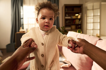 Baby standing with support from Black adult hands in living room, baby looking forward with curious...