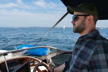 A bearded man in sunglasses and a cap enjoys his summer vacation, confidently driving a motorboat...