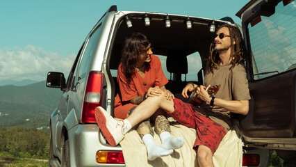 Happy couple relaxing in car trunk with mountain view, playing ukulele and enjoying road trip moments together. Carefree lifestyle and love.