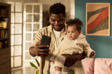 Black young adult man holding infant child while taking selfie with smartphone in home setting,...