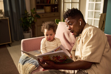 Black young adult man sitting on sofa reading picture book to child, both engaging with story, man...