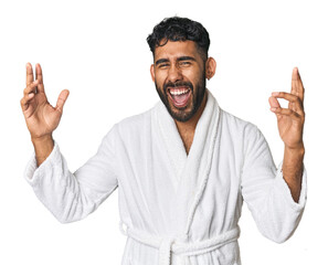Hispanic male in a robe in studio receiving a pleasant surprise, excited and raising hands.