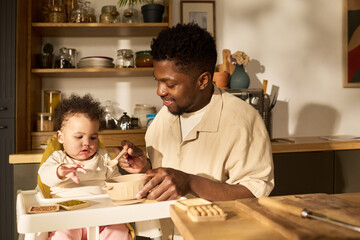 Black young adult man feeding baby girl in high chair during daytime in kitchen, father smiling...