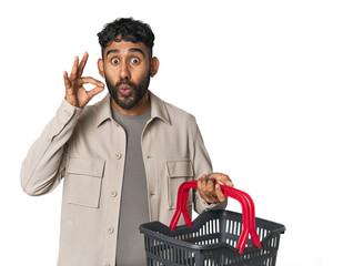 Hispanic male holding shopping basket in studio with fingers on lips keeping a secret.