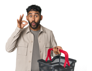 Hispanic male holding shopping basket in studio with fingers on lips keeping a secret.