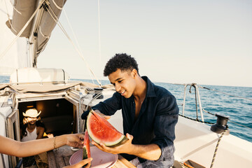 Sailor offering watermelon slice to friends on sailboat during summer vacation, enjoying refreshing...