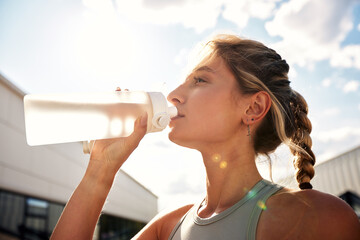 Slender female athlete in gray sportswear with a sports bottle of water against the summer blue sky...