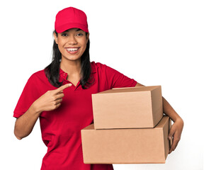 Young Filipina courier with two packages smiling and pointing aside, showing something at blank...