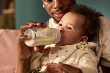 Black man feeding infant with bottle while holding baby in arms, demonstrating paternity leave concept, close up showing father bonding with young child at home