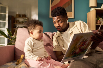 Black young adult man sitting on sofa reading picture book to toddler child during paternity leave,...