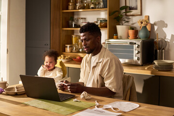Baby sitting beside Black young adult man working on laptop at kitchen table, man holding smartphone and looking at screen, baby smiling, paternity leave concept