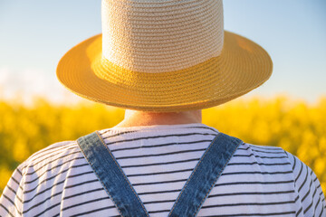 Rear view of female farmer with straw hat standing in blooming rapeseed field and looking into...