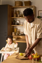 Black young adult man preparing fruit at kitchen counter while infant child sitting in high chair...