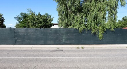 Green tarpaulin on vinyl coated fence at the roadside. Trees,  rooftops and sky on behind, sidewalk...