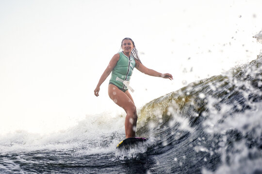 Young beautiful sporty girl in a green life jacket surfing on the wave. Happy sportswoman showily glides on the board. Active lifestyle, healthy hobby. Film grain effect.