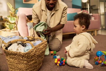 Black man kneeling on floor caring for infant child during paternity leave, folding laundry while...