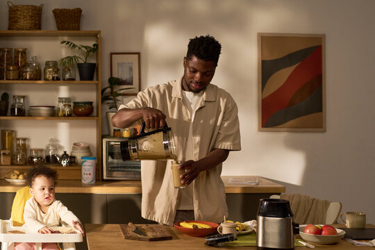 Black young adult man preparing smoothie in kitchen while caring for child sitting at table, father multitasking during paternity leave, morning domestic routine