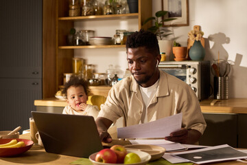 Black young adult man working on laptop and holding documents while sitting at kitchen table with...