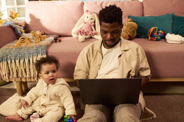 Black young adult man sitting on floor using laptop while caring for baby child playing with toys...